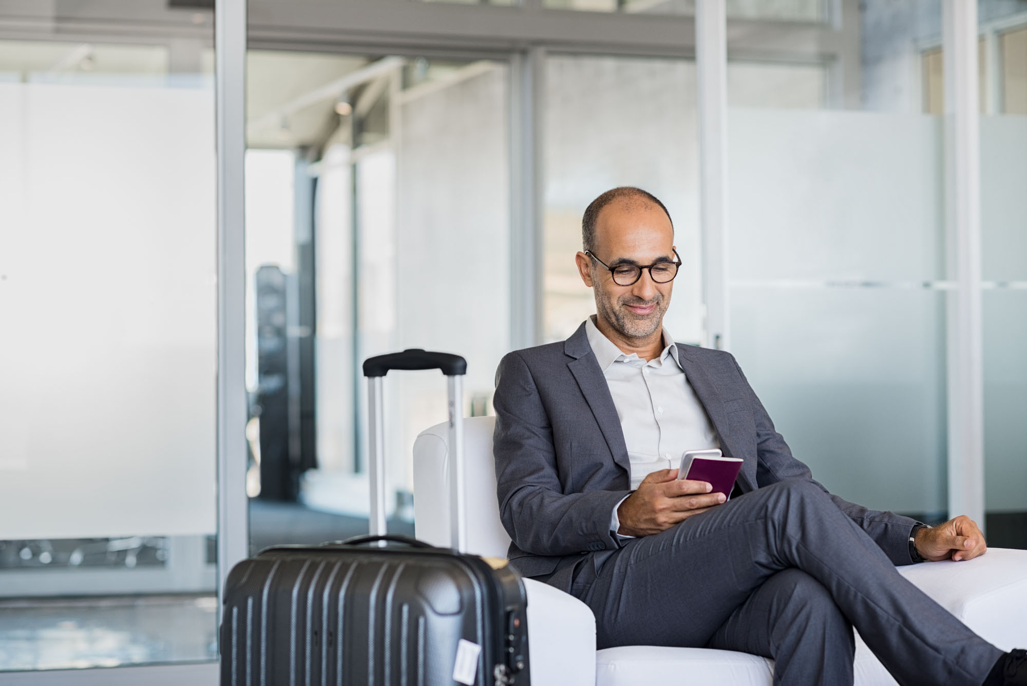 Mature businessman using mobile phone at the airport in the waiting room. Business man typing on smartphone in lounge area. Portrait of latin man sitting and holding passport with luggage.
